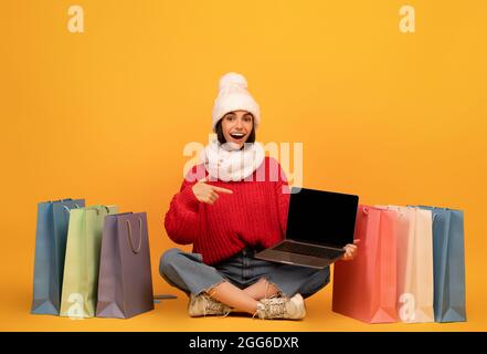 Bonne femme shopaholic en tenue d'hiver pointant à l'écran d'ordinateur portable, assis parmi les sacs de cabas, fond jaune Banque D'Images