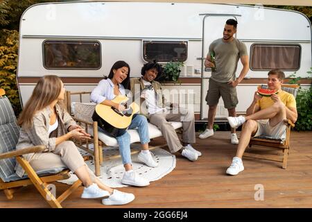 Divers amis jouant de la guitare et du chant, buvant de la bière, mangeant de la pastèque, assis dans des chaises longues lors d'un voyage de camping Banque D'Images