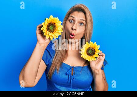 Femme hispanique d'âge moyen tenant des tournesols jaunes dans le visage de choc, regardant sceptique et sarcastique, surpris par la bouche ouverte Banque D'Images