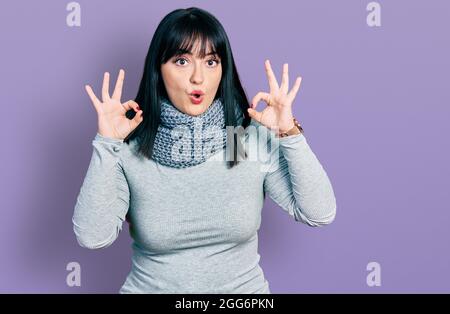 Jeune femme hispanique de taille plus portant un foulard d'hiver regardant surpris et choqué faisant ok symbole d'approbation avec les doigts. Expression folle Banque D'Images