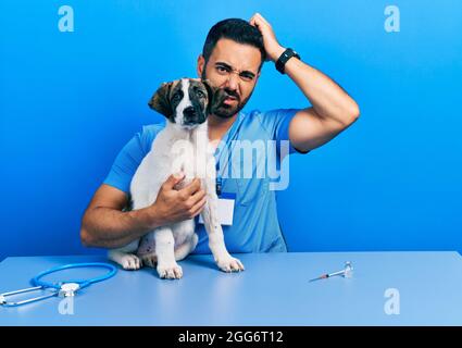 Beau vétérinaire hispanique avec la barbe de vérifier la santé de chien confondre et se poser des questions. Incertain avec le doute, penser avec la main sur la tête Banque D'Images