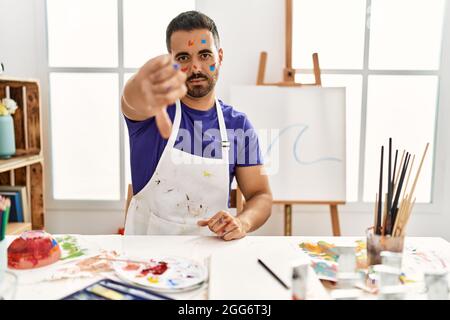 Jeune homme hispanique avec la barbe dans un studio d'art avec le visage peint qui a l'air malheureux et en colère montrant le rejet et négatif avec le pouce vers le bas geste. Mauvais e Banque D'Images