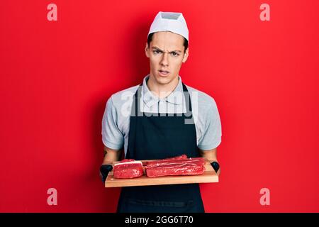 Un jeune hispanique tient une planche avec de la viande crue dans le visage choqué, regardant sceptique et sarcastique, surpris par la bouche ouverte Banque D'Images