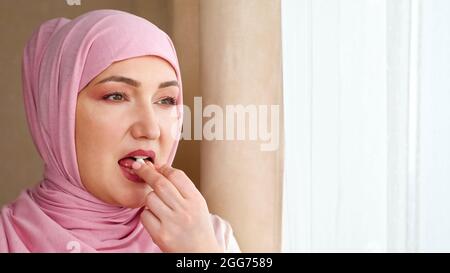 Une jeune femme en hijab boit une capsule blanche avec de l'eau tout en se tenant près de la fenêtre Banque D'Images