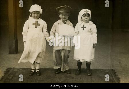 Vers 1917, historique, trois petits enfants debout ensemble vêtus de costumes habillés de fantaisie, les deux petites filles comme infirmières, le petit garçon comme un officier blessé de l'armée avec son bras gauche dans une élingue, Angleterre, Royaume-Uni. Banque D'Images