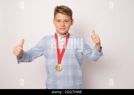 Jeune garçon excité montrant sa médaille d'or avec les pouces vers le haut sur fond blanc. Banque D'Images