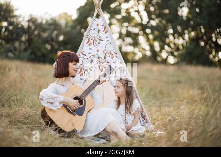 Jolie jeune mère et fille jouant de la guitare ensemble, assis à côté de la tente de tipi à l'extérieur dans le champ d'été. Boho femme indienne jouant de la guitare pour sa mignonne fille heureuse enfant Banque D'Images