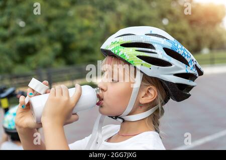 Mignon petite fille caucasienne enfant dans casque de sport de sécurité boire de l'eau fraîche de la bouteille après l'exercice vélo, scooter ou patins à roulettes sur le parc de la ville Banque D'Images