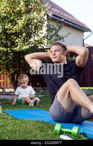 Jeune homme avec enfant va dans pour le sport à la maison dans l'arrière-cour en été, formation en ligne. L'athlète fait la presse sur le tapis dans le jardin, sur b Banque D'Images