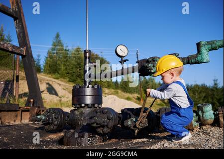 Adorable petit garçon en casque de construction à l'aide d'une clé industrielle lors de la réparation du tuyau du cric de pompe de puits d'huile. Un ouvrier de l'huile mignon serre le boulon sur le tuyau lorsqu'il travaille dans le champ d'huile. Banque D'Images
