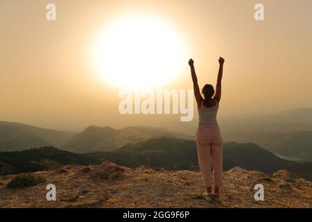 Vue arrière portrait d'une seule femme levant les bras au coucher du soleil célébrant dans la montagne Banque D'Images