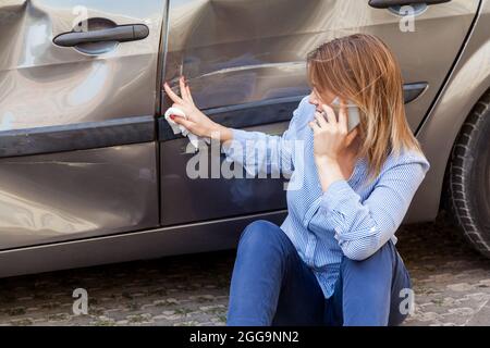 Blonde femme dans une tenue décontractée assis près de la voiture bosselée et touchant les rayures et les bosses sur la porte d'automobile, parlant téléphone avec l'agent d'assurance ou demander Banque D'Images