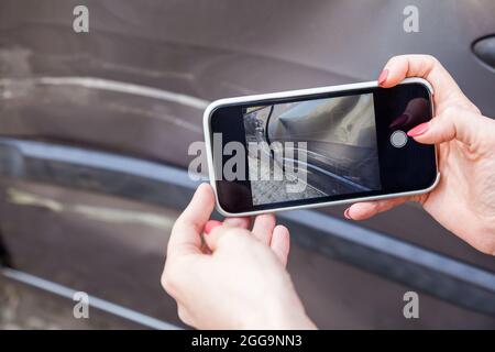 Gros plan des mains des femmes avec un smartphone qui photographie les conséquences de l'accident de la route, des rayures et des bosses sur l'automobile, la partie latérale endommagée de la voiture Banque D'Images
