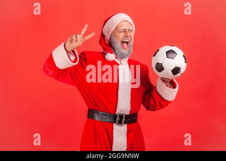 Homme âgé stupéfié avec une barbe grise portant le costume du père noël tenant le ballon de football dans les mains, montrant le signe v à l'appareil photo, en se vrillant, bouche ouverte. stu. Intérieur Banque D'Images
