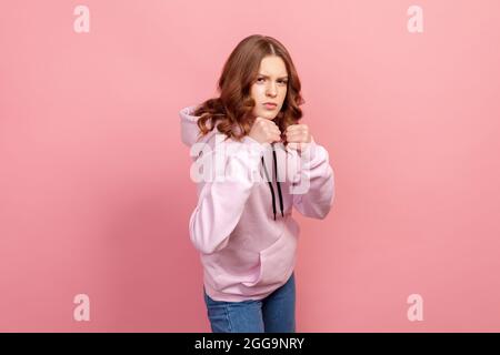 Portrait d'une adolescente en colère à capuche debout avec des poings serrés et un look furieux, prêt à punch. Prise de vue en studio, isolée sur fond rose Banque D'Images