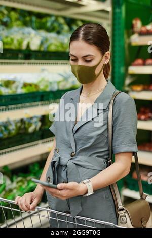 Jeune femme enceinte en masque facial et robe debout au panier dans le département des légumes du supermarché et liste de contrôle sur le téléphone Banque D'Images