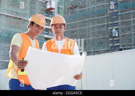 Équipe de travailleurs de la construction discutant des détails du plan de construction lorsqu'ils se trouvent sur le site avec des gratte-ciel en arrière-plan Banque D'Images