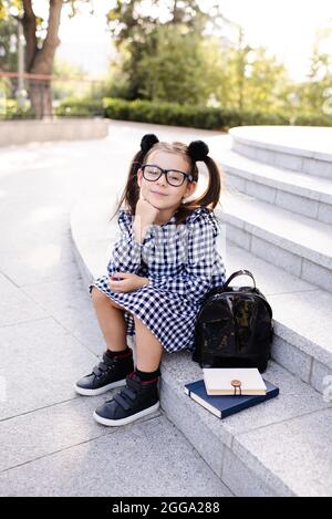 Sourire heureux enfant fille 5-6 ans porter uniforme robe et des lunettes assis sur l'escalier avec des livres et sac à dos dehors. Regarder l'appareil photo. Retour à la goélette Banque D'Images