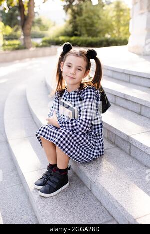 Mignon jeune fille 5-6 ans vêtements à carreaux noir et blanc robe et sac à dos tenant des livres assis sur les escaliers à l'extérieur de près. Regarder la came Banque D'Images