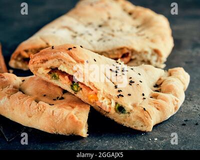 Parfait calzone de pizza fermée maison sur des feuilles de papier de cuisson sur fond noir. Couper de savoureuses pizzas italiennes fermées avec du fromage, de la viande, des légumes, du sésame noir habillé. Espace de copie pour le texte Banque D'Images