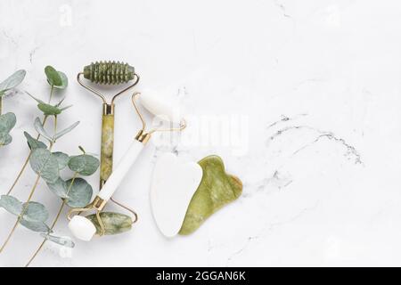 Outils de massage. Masseur Gua Sha, rouleaux faciaux jade et branches d'eucalyptus sur fond de marbre blanc. Concept SPA. Vue de dessus, espace de copie Banque D'Images