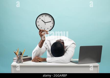 Homme d'affaires afro-américain fatigué assis à un bureau avec un ordinateur portable, tenant une horloge, se sentant épuisé, fond bleu Banque D'Images