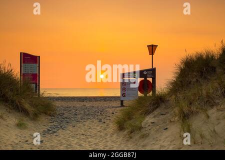 Vue entre ces dunes de sable du Royaume-Uni en regardant vers un coucher de soleil sur la mer avec des panneaux d'avertissement de plage. Banque D'Images