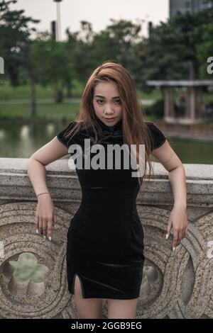 Belle jeune femme asiatique dans une robe noire décontractée debout près de la main courante dans le parc. Cheveux longs. Bonne posture. Jolie femme vietnamienne. Banque D'Images
