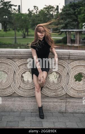 Belle jeune femme asiatique dans une robe noire décontractée debout près de la main courante dans le parc. Vent dans ses cheveux. Bonne posture. Jolie femme vietnamienne. Banque D'Images