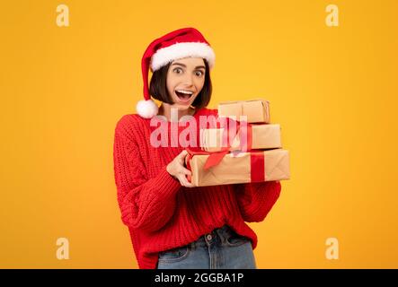 Femme excitée en tenue d'hiver et chapeau de père noël tenant des boîtes-cadeaux sur fond jaune studio, espace de copie Banque D'Images