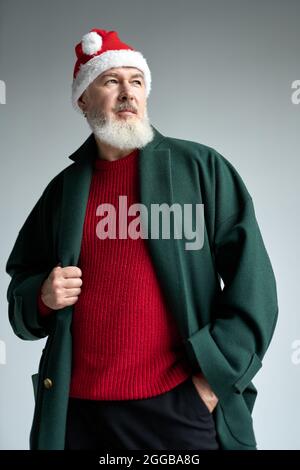 Cool homme d'âge moyen avec chapeau de Noël portant une tenue élégante regarder loin, en ajustant son manteau tout en posant isolé sur fond gris clair Banque D'Images