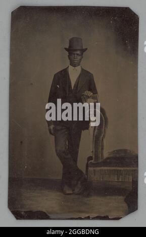 Sans titre (Portrait d'un homme debout), 1875. Portrait noir et blanc d'un homme noir rasé debout dans un chapeau et appuyé contre un fauteuil, tenant un livre. Banque D'Images