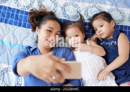 Une jolie jeune femme souriante, couchée au lit avec ses deux petites filles et emportant le selfie sur son smartphone Banque D'Images