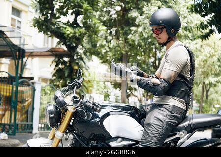 Beau homme sérieux assis sur la moto et mettant des gants en cuir et un casque pour la protection Banque D'Images