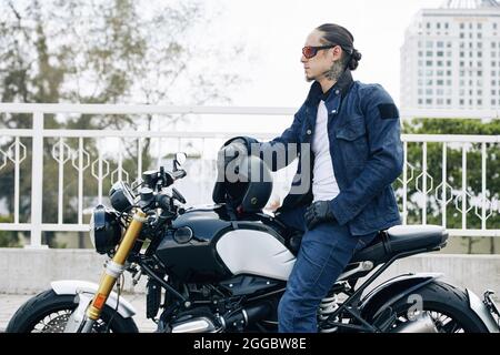 Beau jeune homme élégant en vêtements denim assis sur une moto avec un casque dans les mains Banque D'Images