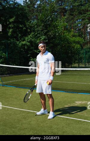 Jeune sportif en vêtements de sport blancs tenant le ballon de tennis et la raquette sur le court Banque D'Images