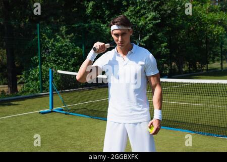 Jeune sportif en vêtements de sport blancs tenant une raquette et un ballon sur un court de tennis Banque D'Images