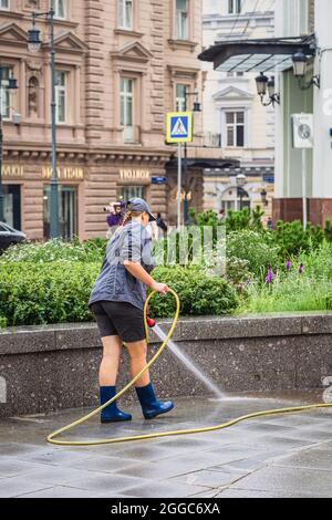 Vue arrière d'une fille méconnue du service de ville arroser des fleurs dans un lit de fleurs et rafraîchir la chaussée par une chaude journée d'été. Les gens ordinaires en public Banque D'Images