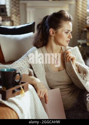 femme moderne pensive en tricoté gilet confortable avec livre, plateau et mug dans le salon moderne en automne ensoleillé. Banque D'Images