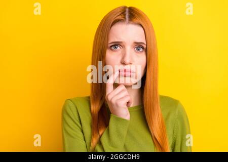 Photo de triste cheveux rouge jeune femme doigt menton porter un chandail vert isolé sur fond jaune Banque D'Images