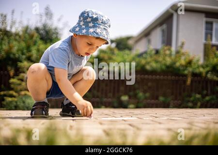 Joyeux petit garçon dessin hopscotch avec craie sur terre appréciant enfance heureuse à l'arrière-cour Banque D'Images