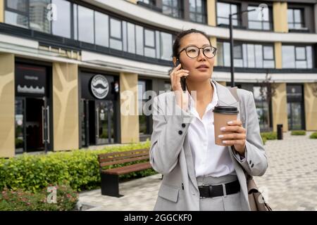 Jeune femme d'affaires confiante avec un smartphone à l'oreille prenant un verre à l'extérieur Banque D'Images
