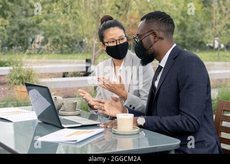 Deux cols blancs en confiance dans les vêtements de coffrage et les masques de protection discutant de nouveau projet d'affaires dans un café extérieur Banque D'Images