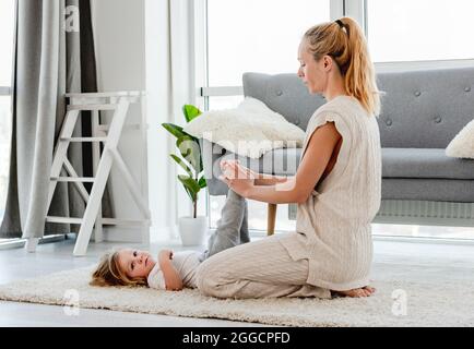 Jeune mère blonde faisant des exercices pour son enfant fils jambes sur le sol à la maison Banque D'Images
