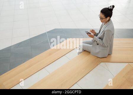 Jeune femme asiatique en costume élégant écoutant le son dans un smartphone tout en étant assise sur de grands escaliers Banque D'Images