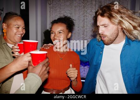 Divers groupes de jeunes amis qui profitent de la fête à l'intérieur et tiennent des tasses rouges, prises avec flash Banque D'Images
