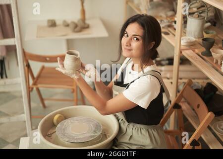 Vue latérale en grand angle d'une jeune femme souriante en tablier, assise à la roue de poterie et démonstration de la verseuse en argile faite à la main pendant la leçon d'art créatif studi Banque D'Images