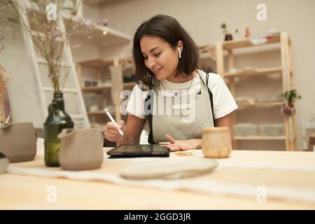 Jeune femme souriante maître en céramique écoutant de la musique grâce aux oreillettes TWS tout en concevant de la vaisselle sur une tablette graphique avec stylet Banque D'Images