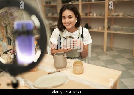 Jeune femme positive souriant et démontrant pichet d'argile à vlog public tout en filant la vidéo et travaillant dans la poterie Banque D'Images