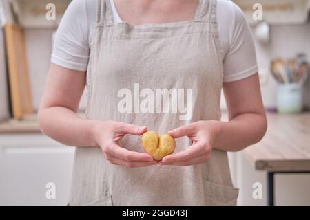 Les mains des femmes tiennent les pommes de terre pelées. Pommes de terre en forme de coeur dans les mains d'une femme dans la cuisine Banque D'Images